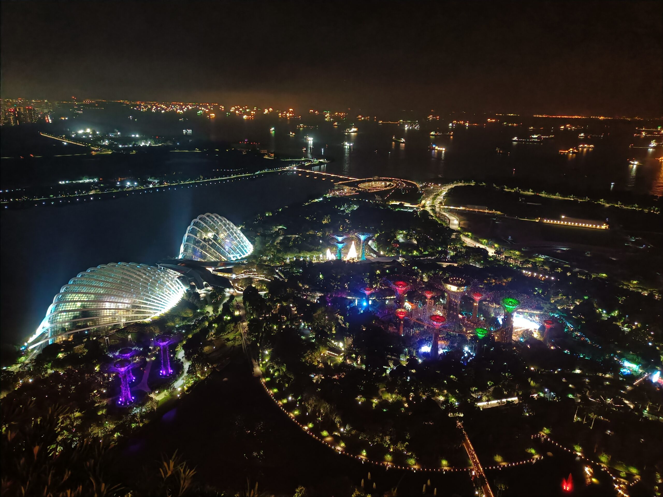 Aerial view of illuminated gardens and waterfront at night with colorful lights and city skyline in the background.