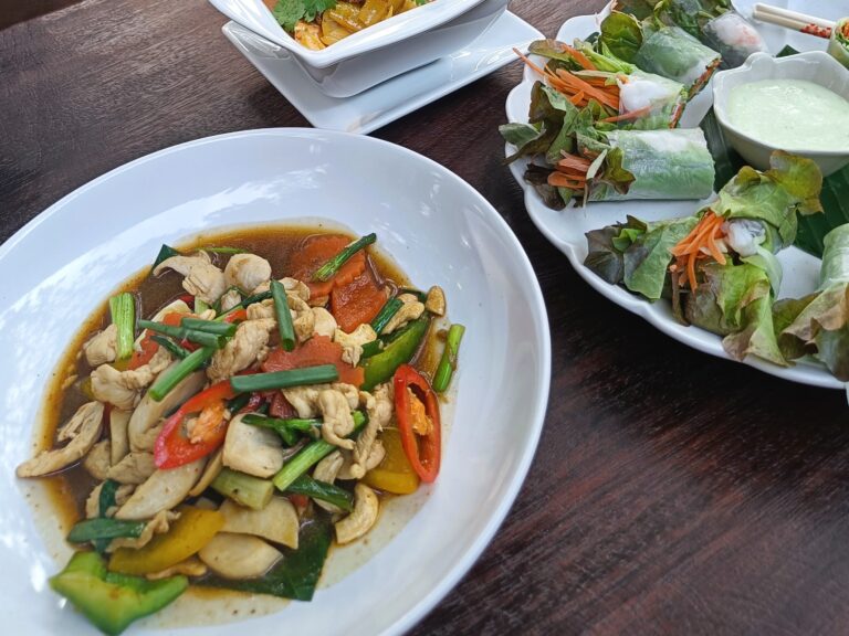 A table with must-try food in Singapore features stir-fried chicken and vegetables, fresh spring rolls, and a creamy dipping sauce.