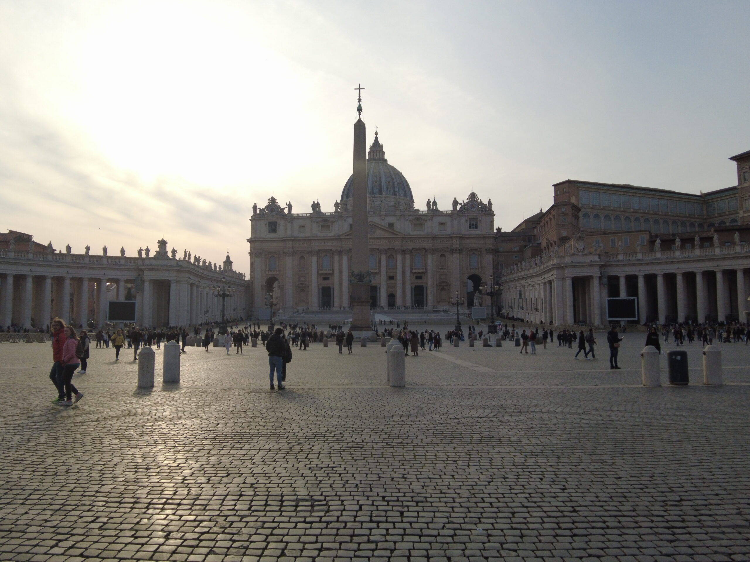 St. Peter's Square, one of the must-see attractions in Rome, bustles with people under a bright sky, the Basilica standing majestically in the background.