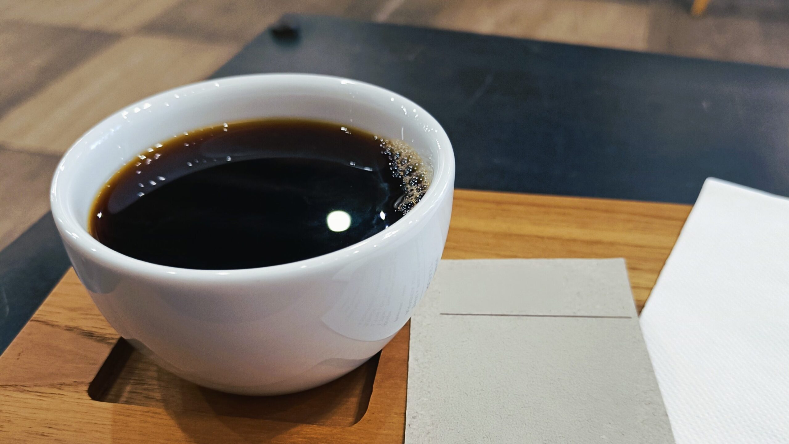 A cup of steaming Kopi rests on a wooden tray with a napkin, elegantly placed on a sleek black table.