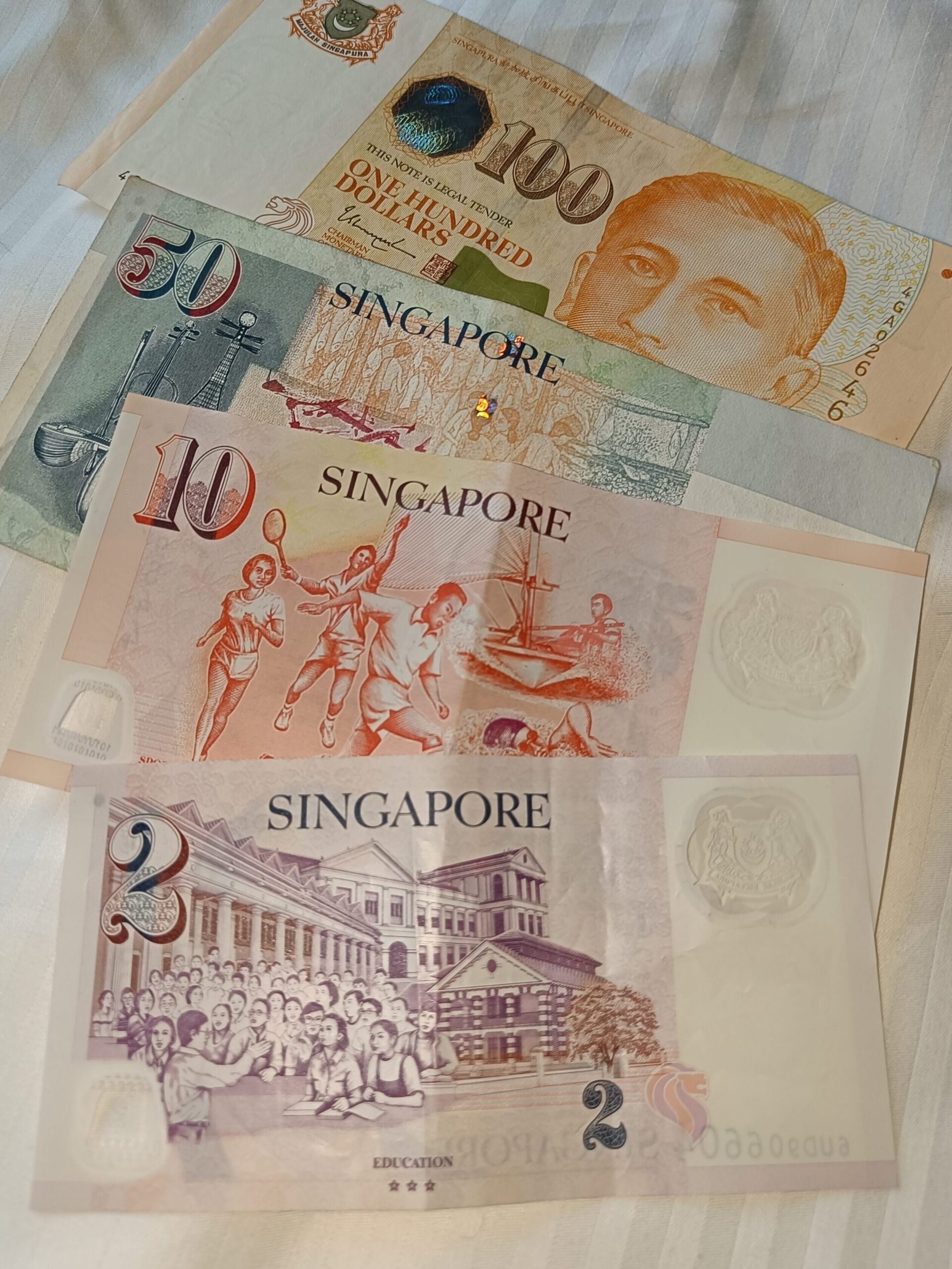 For a first-time visit to Singapore, familiarize yourself with the local currency: Singapore dollar banknotes in denominations of 100, 50, 10, and 2. Here they are beautifully arranged on a white fabric background.