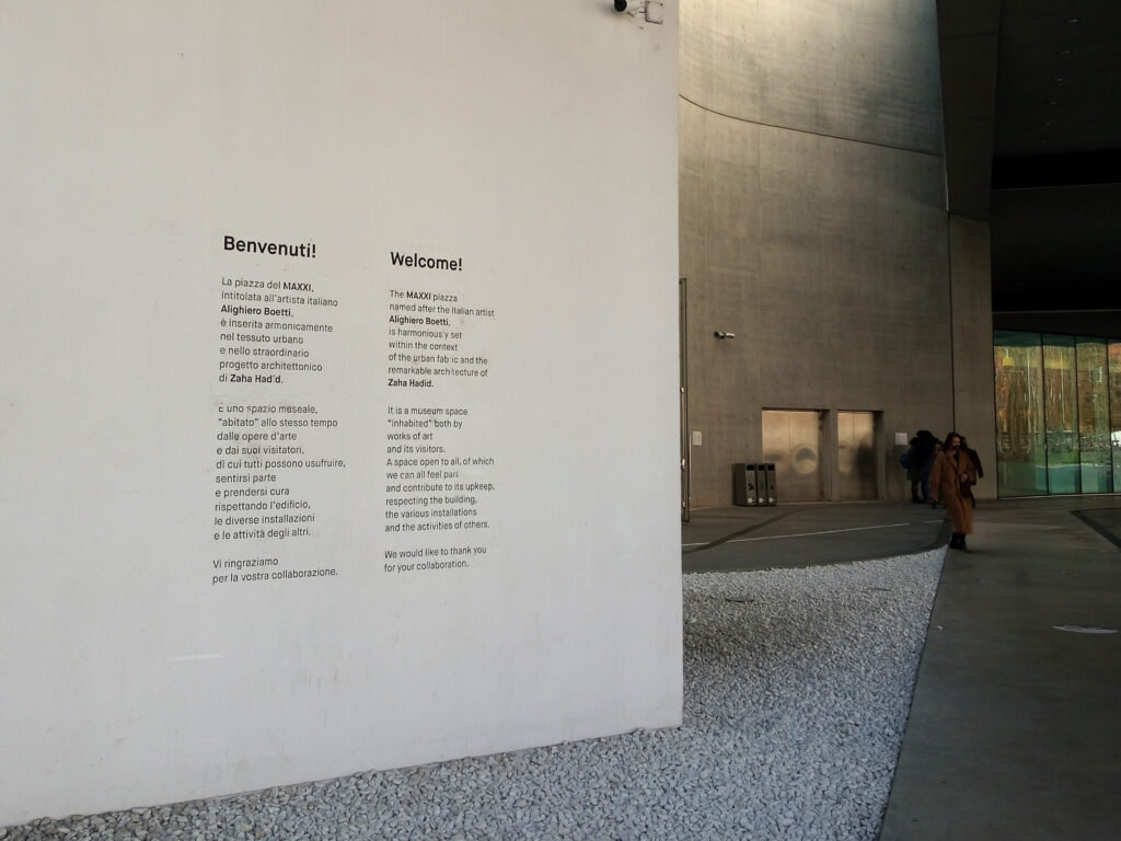 Modern building entrance with a welcome sign on the wall; person walking toward glass doors; minimalist design reminiscent of the MAXXI museum in Rome.