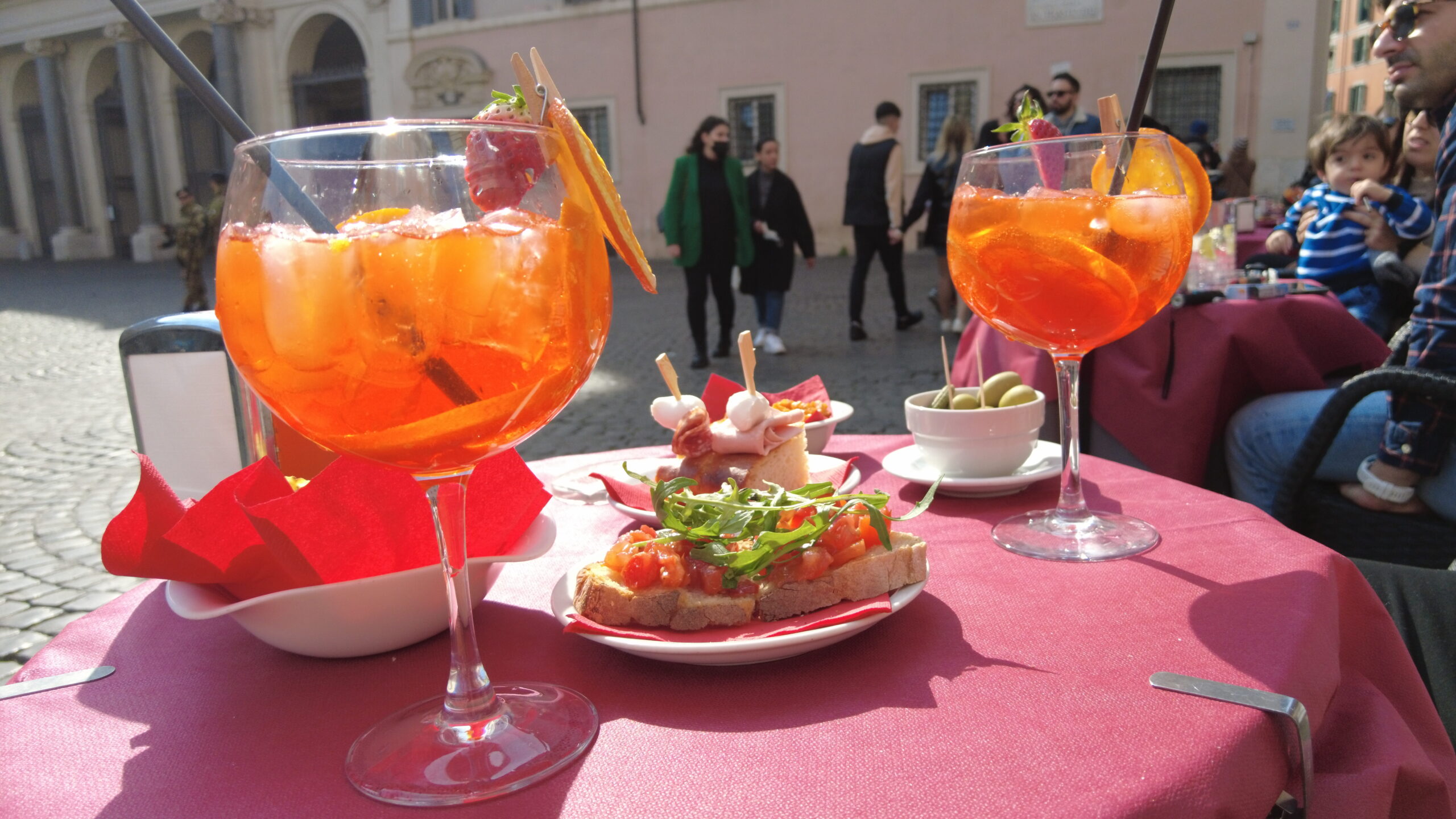 Two glasses of orange cocktails sit on a table with appetizers, capturing the lively vibe of a popular outdoor café in one of Rome's charming neighborhoods.