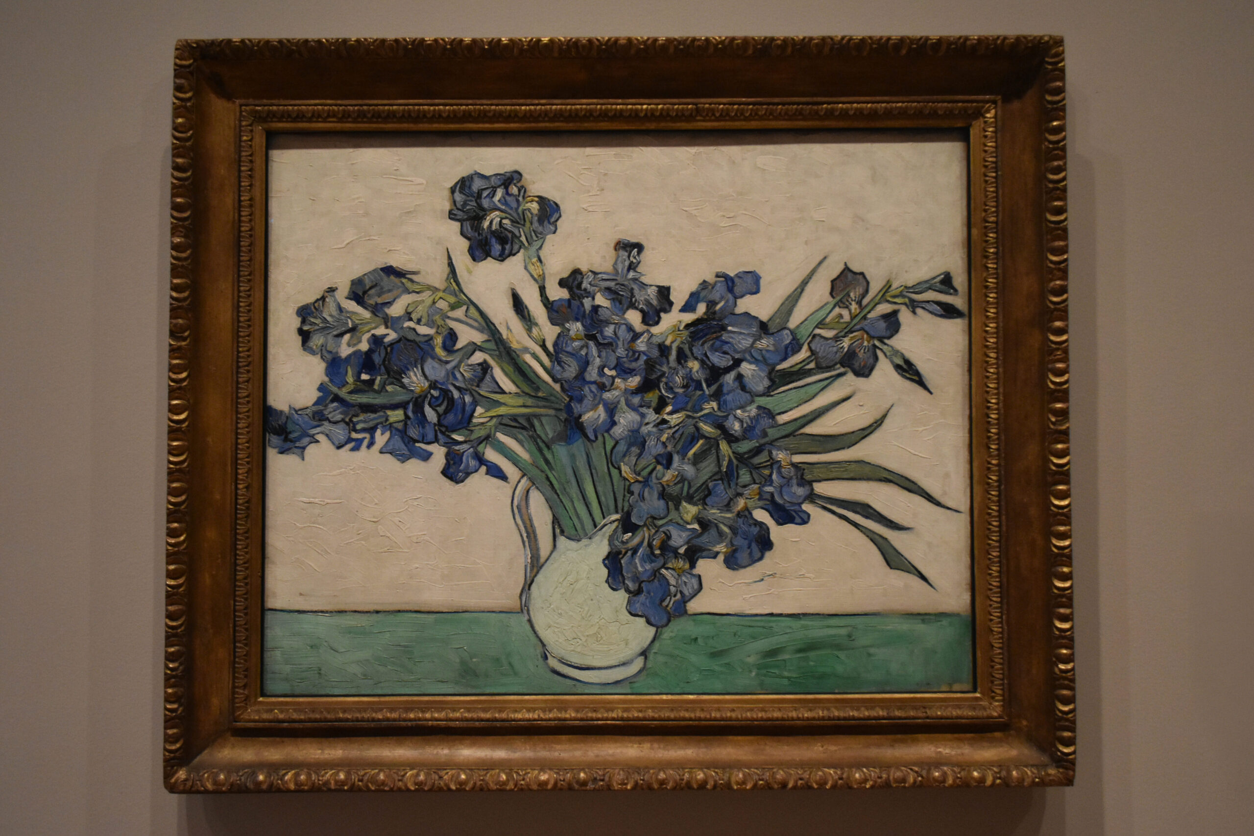 Framed painting of blue irises in a vase against a beige background, reminiscent of the vibrant beauty captured on a Van Gogh tour through Provence, displayed on a gray wall.