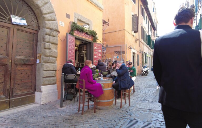 People seated outdoors at a café on a cobblestone street in one of Rome's popular neighborhoods, with colorful buildings in the background.