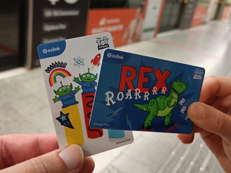 Two hands holding EZ-Link cards featuring Toy Story characters—one with aliens and one with Rex the dinosaur. A must-have for any first-time visitor, these cards are not just collectibles but your ticket to smooth travel. Don't forget to add them to your Singapore guide essentials!.