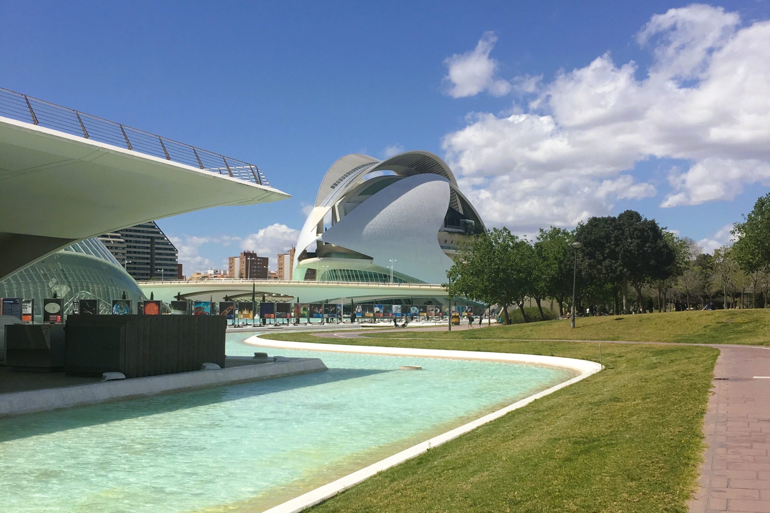 Modern architectural buildings in Valencia line a canal, with trees swaying under a blue sky with clouds. This scene captures the essence of one of the city's popular neighborhoods.