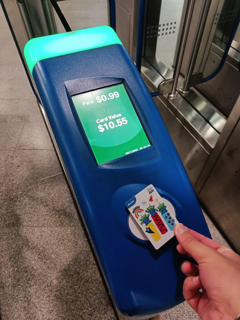 Person tapping a transit card on a blue terminal displaying fare and card balance at a station during their first-time visit to Singapore, where essential tips for navigating public transport make travel smooth and efficient.