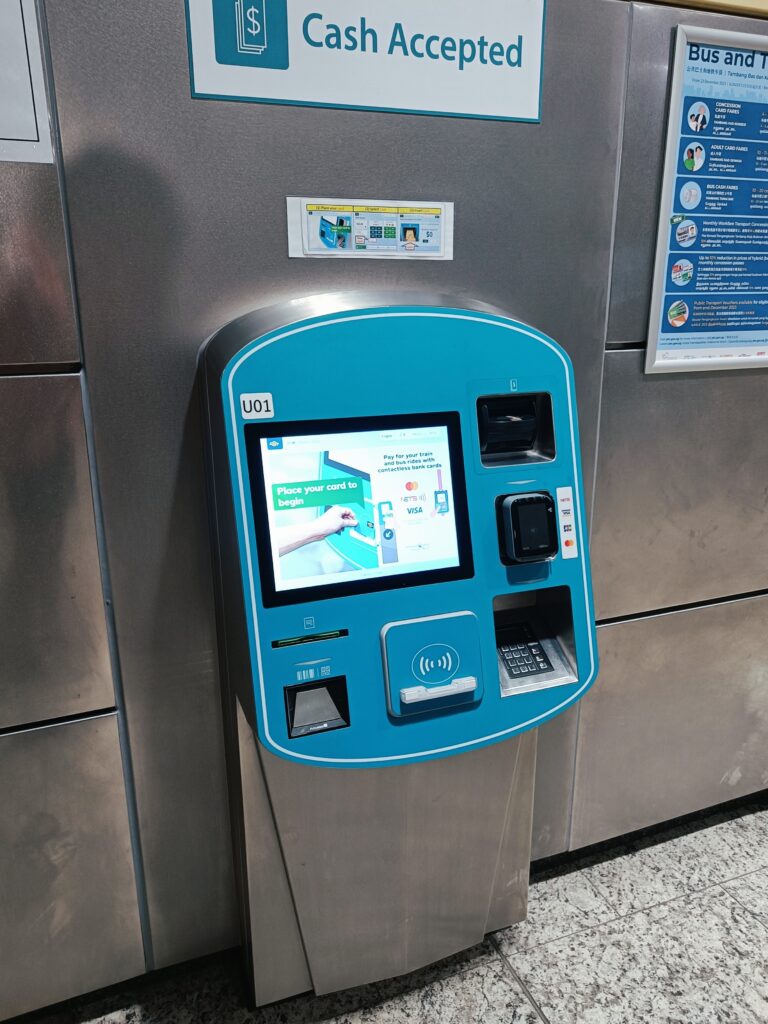 Ticket machine with a touchscreen and card payment option, placed against a metal wall with informational signs—essential tips for first-time visitors to Singapore looking to navigate the city's efficient transport system.