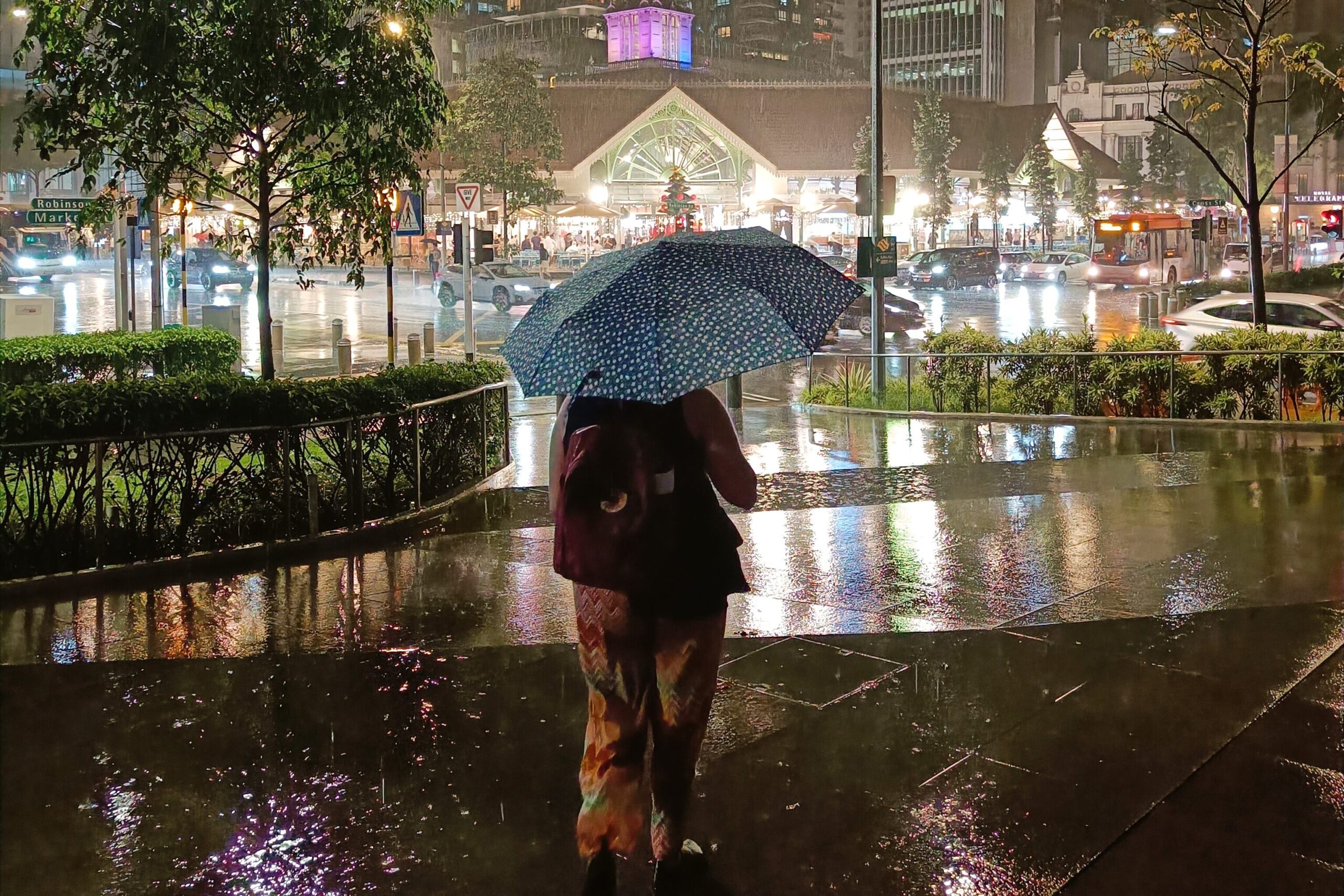 A person with a blue umbrella explores the rain-soaked streets of Singapore at night, surrounded by towering buildings and vibrant lights. For a first-time visit, remember to embrace the city's unexpected downpours with enthusiasm—it's one of the best tips for enjoying your stay.