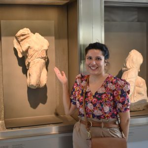 A woman in a floral shirt stands beside ancient sculptures in a museum display.