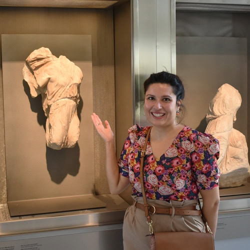 A woman in a floral shirt stands beside ancient sculptures in a museum display.