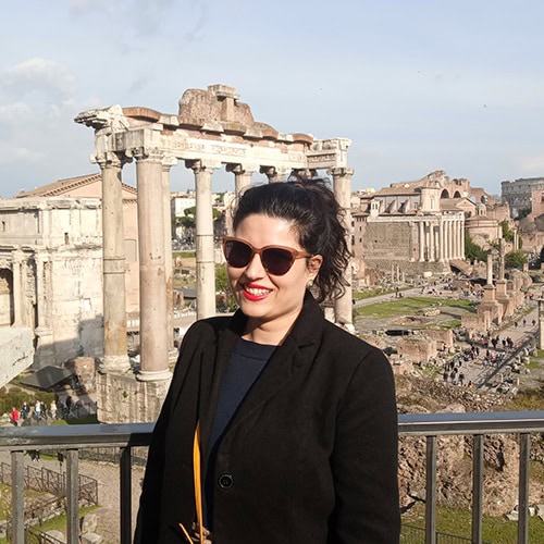 A woman in sunglasses and a black coat stands smiling in front of ancient ruins on a sunny day.