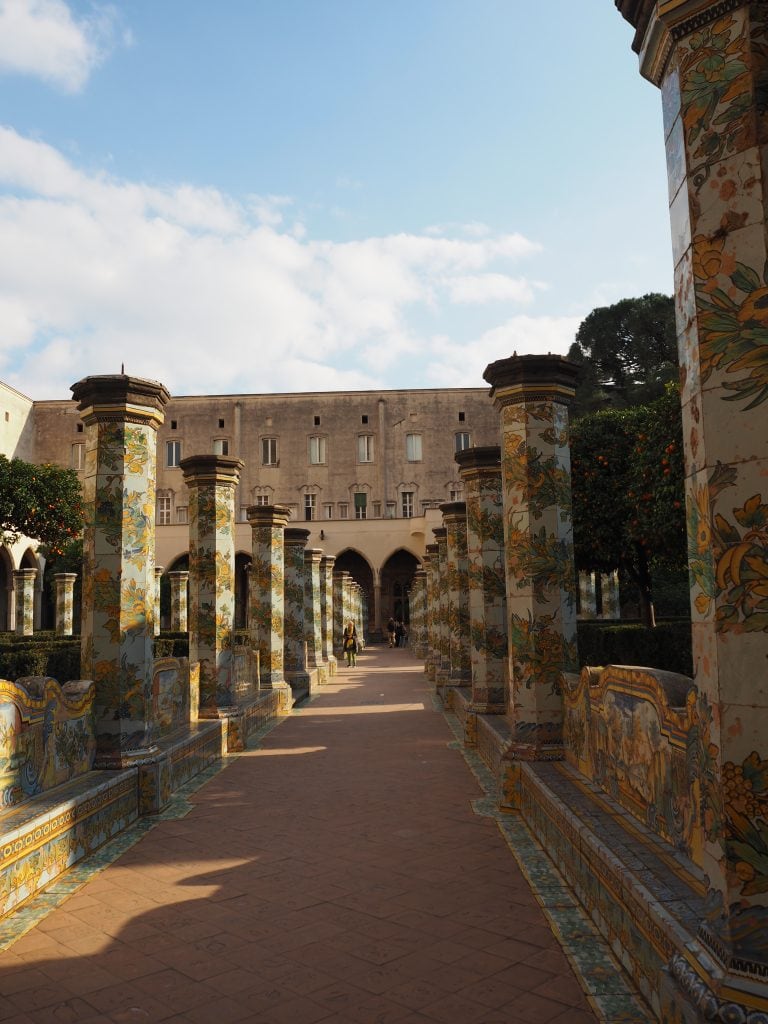 Cloister walkway with ornate columns, set against a historic building under Naples' blue sky—a true highlight among the best things to do in this charming city.