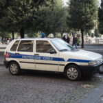 Small white car with "Protezione Civile" parked on a cobblestone street, surrounded by trees.