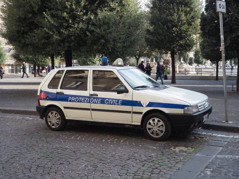 Small white car with "Protezione Civile" parked on a cobblestone street, surrounded by trees.