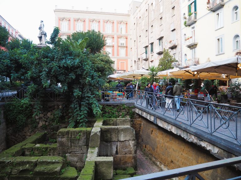 Outdoor cafe with people in one of Naples’ popular neighborhoods, surrounded by ancient stone ruins, lush trees, and tall city buildings in a lively square.