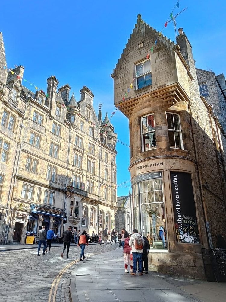 People walking on a sunny street lined with tall historic buildings and colorful bunting overhead, capturing the lively charm of some of the best attractions Edinburgh has to offer.