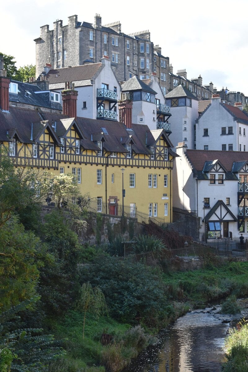 Colorful houses with timber details line a grassy hillside above a small river under a partly cloudy sky, creating a scene reminiscent of some of the best attractions Edinburgh has to offer.
