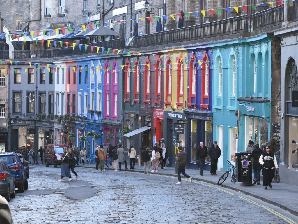 Colorful buildings line a busy cobblestone street with people walking and string flags overhead—a perfect scene for any Edinburgh itinerary.