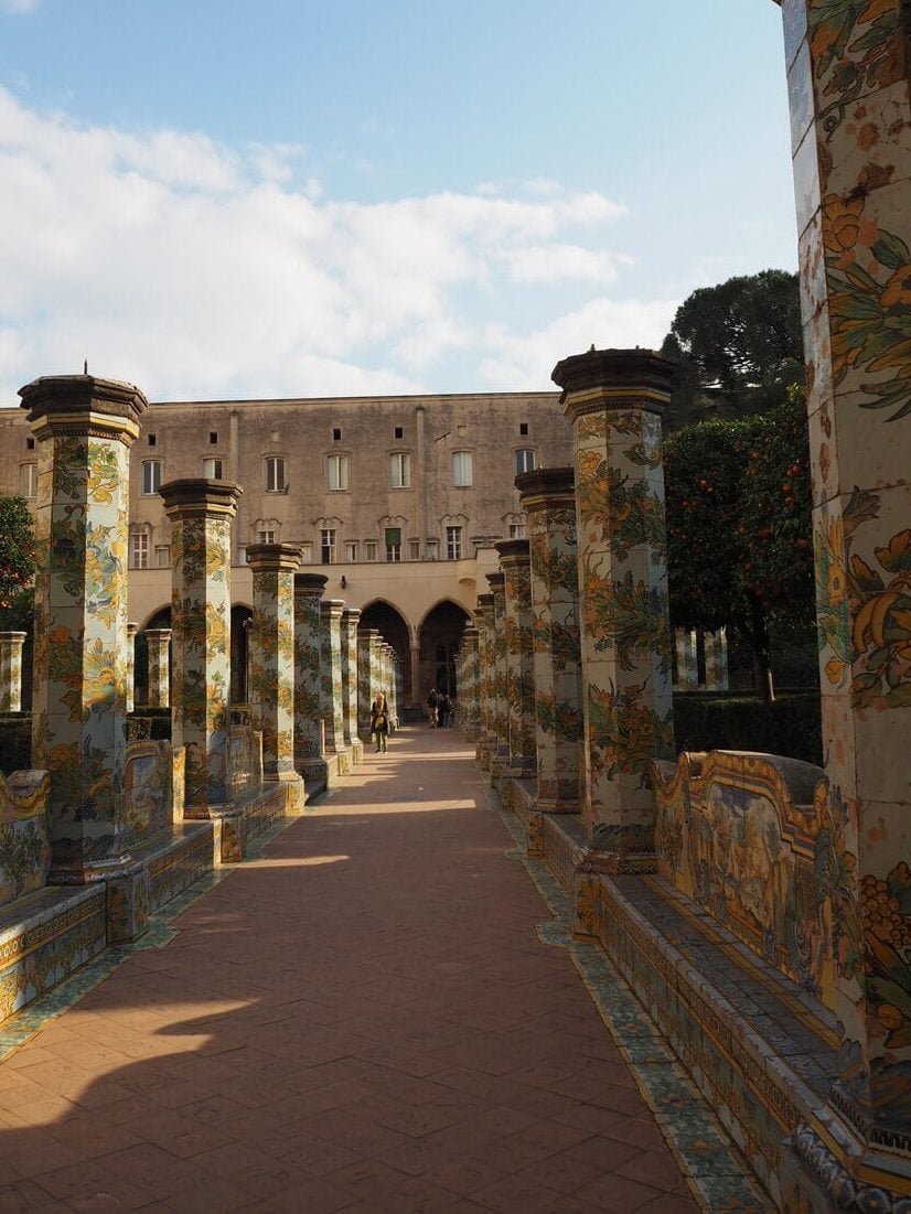 A walkway lined with colorful tiled columns and benches in a historic courtyard under a blue sky—one of the top attractions Naples has to offer.