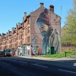 A mural of a bearded man with birds, painted on the side of a tall brick building along a city street.