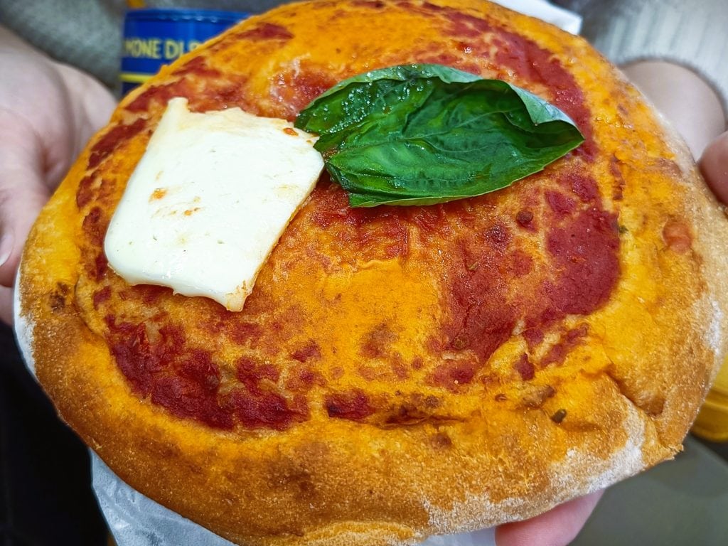 A hand holds a small pizza topped with tomato sauce, basil leaf, and a slice of melted cheese—a delicious sample of must-try food Naples is famous for.
