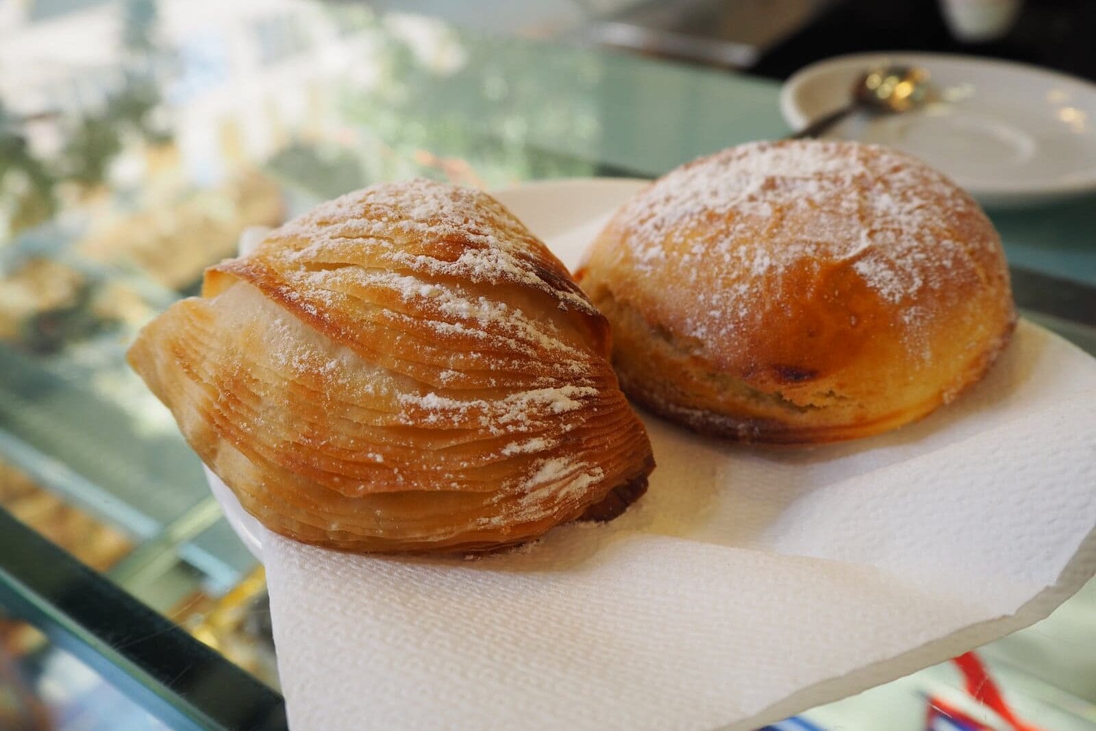 Two Italian pastries with powdered sugar rest on a napkin, displayed on a glass counter—a must try food in Naples and a delicious example of Naples cuisine.