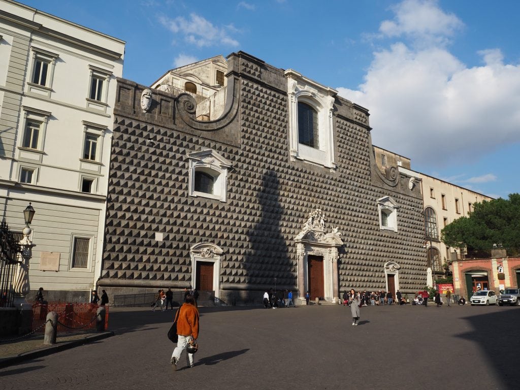 People walk in front of a large, dark stone building with a patterned facade and arched windows under a blue sky, one of the best places in Naples to admire historic architecture.