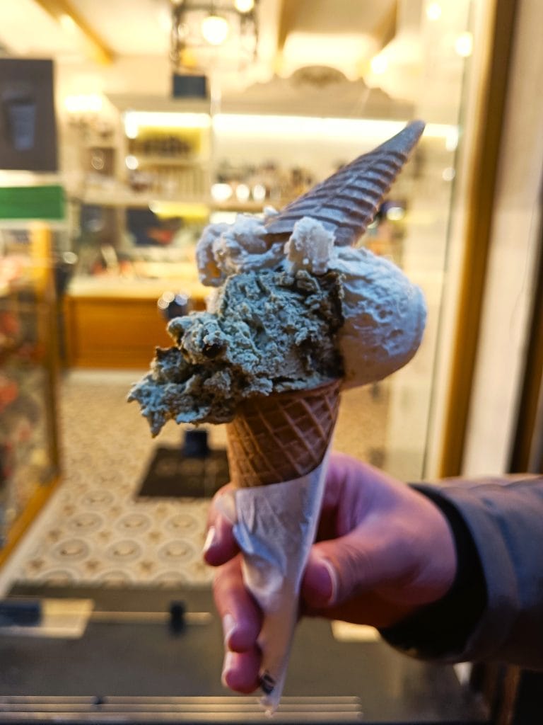 A hand holding a double-scoop ice cream cone with two flavors, in a shop with warm lighting—a delicious treat inspired by must try Naples food.