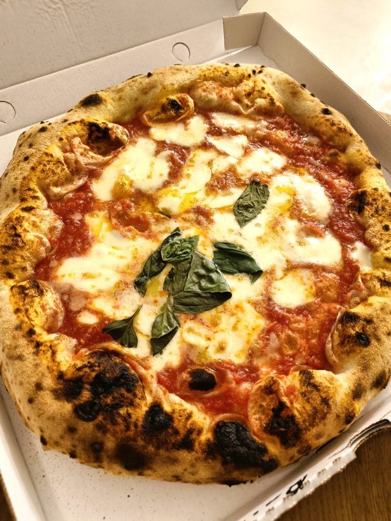 A wood-fired Margherita pizza with melted cheese, tomato sauce, and basil leaves in a white takeout box—a classic must-try Naples food that embodies authentic Naples cuisine.