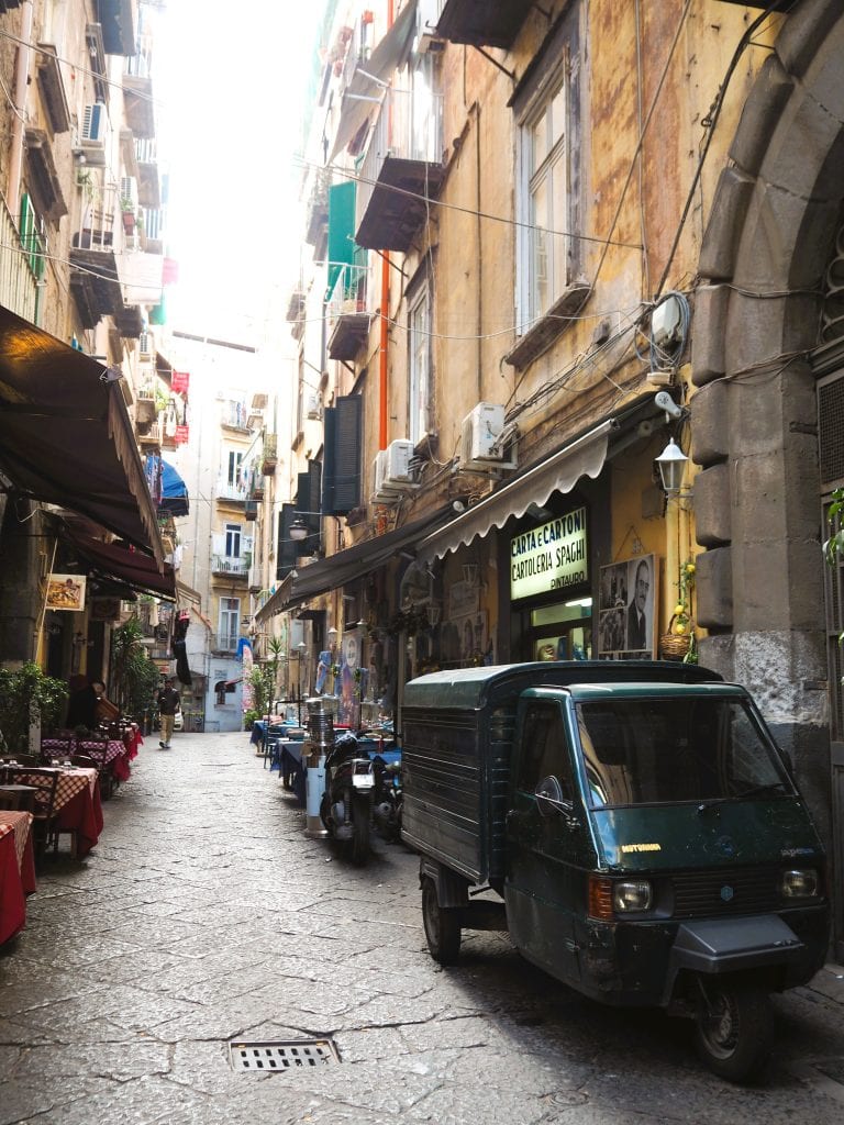 Narrow cobblestone street in one of the popular neighborhoods of Naples, with parked scooters and small vehicles beside outdoor cafes and old buildings, showcasing the lively charm typical of Naples neighborhoods.