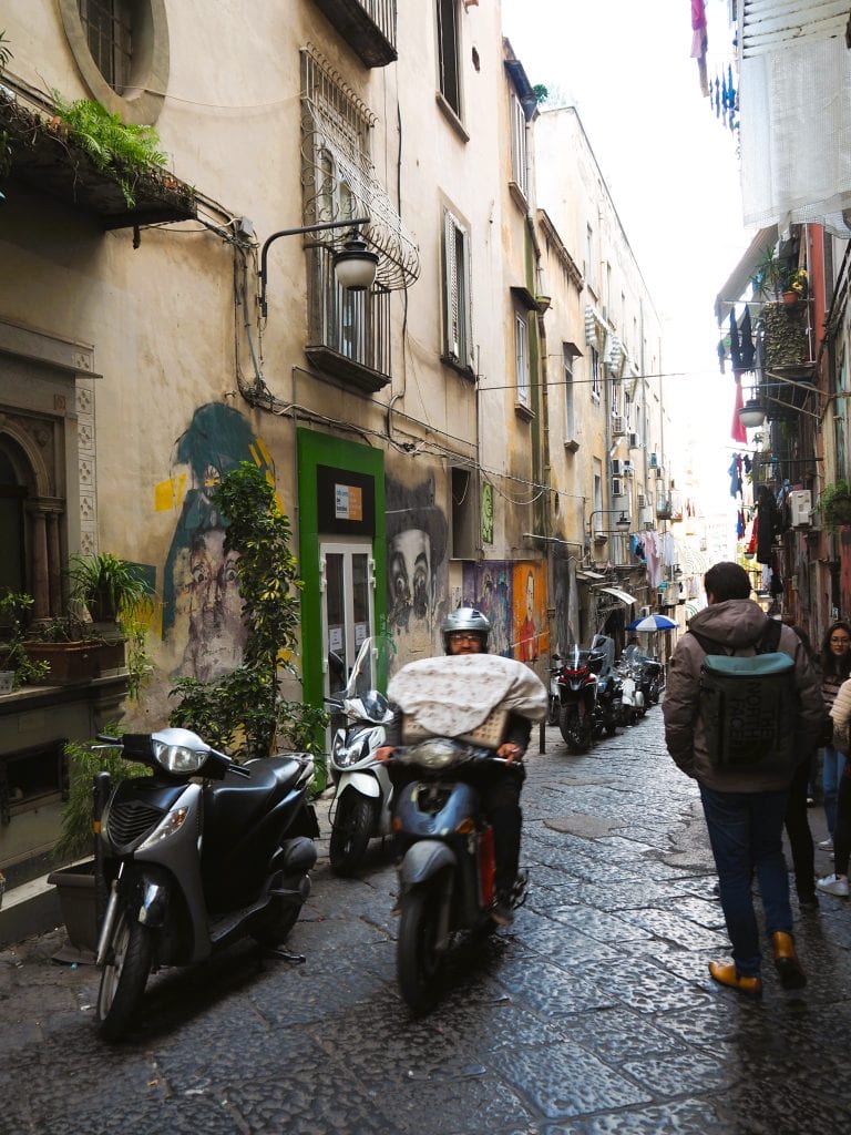 Narrow street in one of the popular neighborhoods of Naples, with scooters, a man riding a scooter, and people walking beside old buildings adorned with murals.