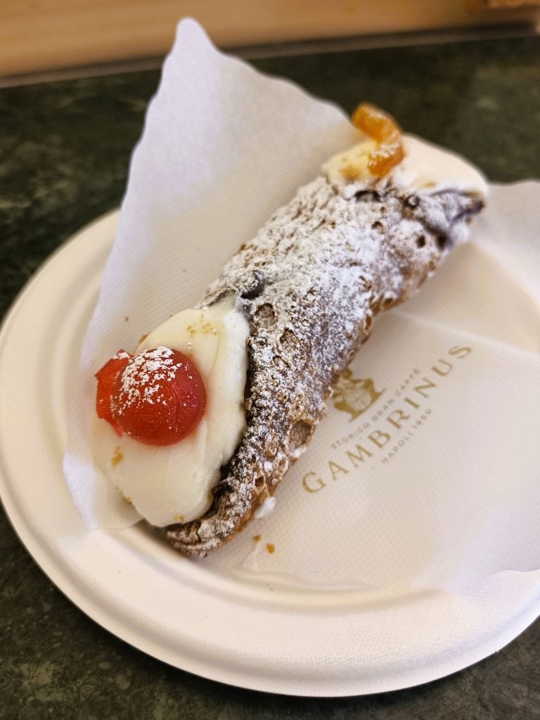 A Sicilian cannolo with ricotta filling, powdered sugar, and candied fruit on a paper plate—a sweet treat often enjoyed alongside other classic Naples cuisine.