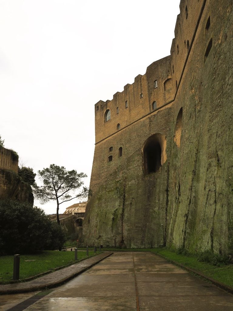 Tall stone fortress walls with small windows rise above a wet pathway, bordered by greenery and trees—a scene reminiscent of the historic charm found in popular neighborhoods of Naples.
