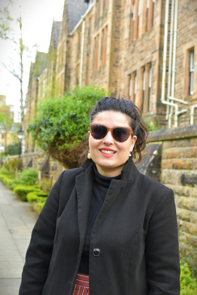 Woman in sunglasses and black coat smiling outside near a stone building and greenery.