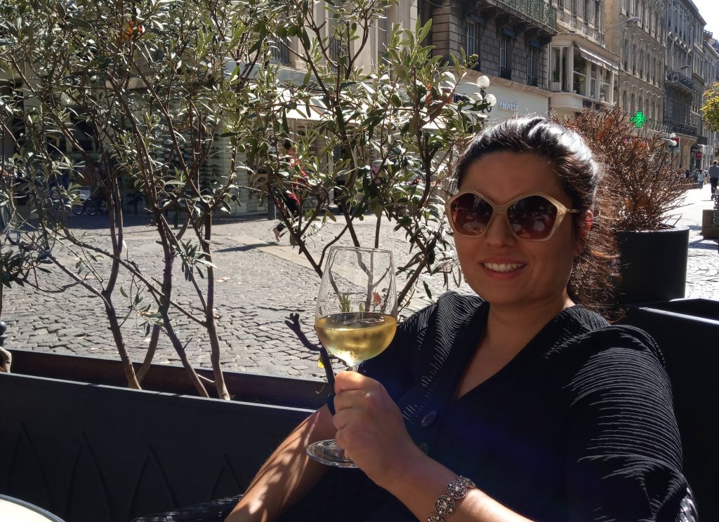 Woman in sunglasses smiling and holding a glass of white wine at an outdoor café on a sunny day.
