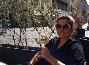 Woman in sunglasses smiling and holding a glass of white wine at an outdoor café on a sunny day.