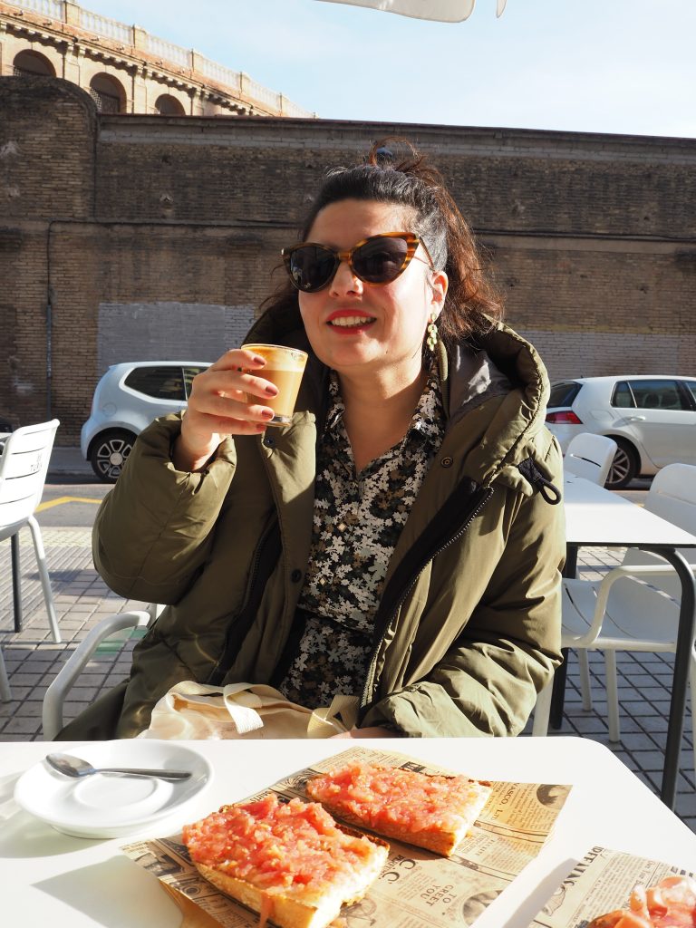 Woman in sunglasses and a green coat drinks coffee at an outdoor café with tomato toast on the table.