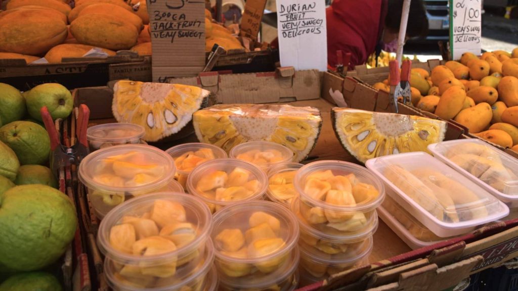 Outdoor fruit market stand with various fresh fruits, including jackfruit, durian, and other tropical delights. Signs show prices for this vibrant assortment of food in New York.