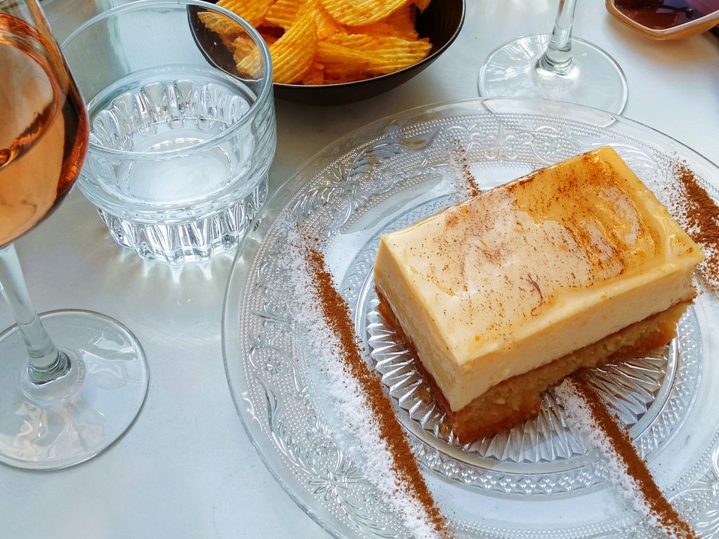 A slice of creamy dessert on a glass plate, with drinks and potato chips nearby on a white table.