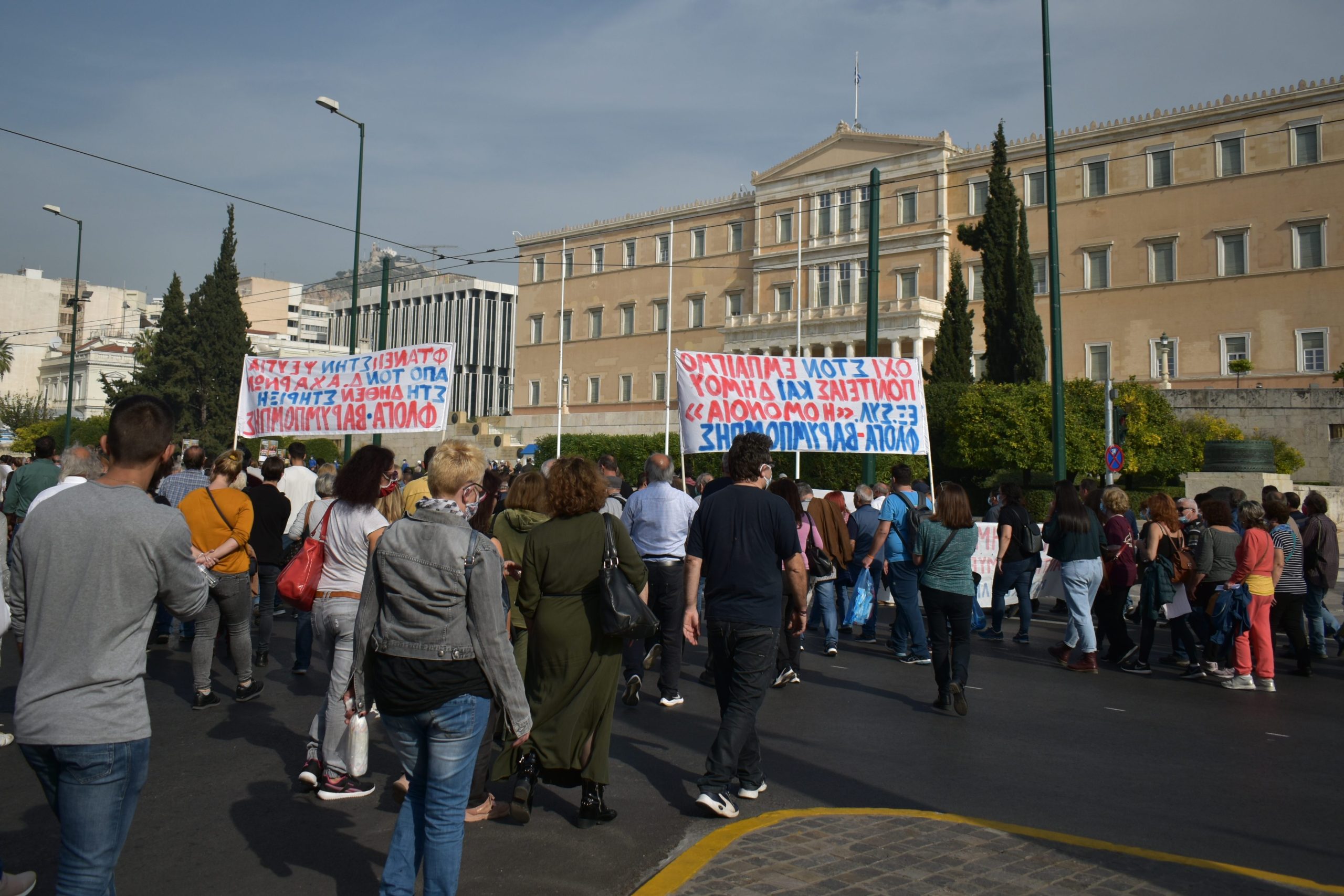 A crowd protests outside a large building in Athens, holding banners with text written in Greek.