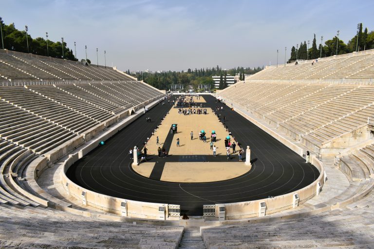Large, empty marble stadium in Athens with a black running track and a few people walking on the field, awaiting crowds for festive dates.