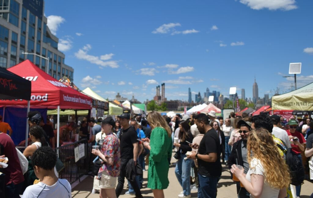 Crowd enjoying food stalls at an outdoor festival on a sunny day with the boroughs of New York City skyline in the background.
