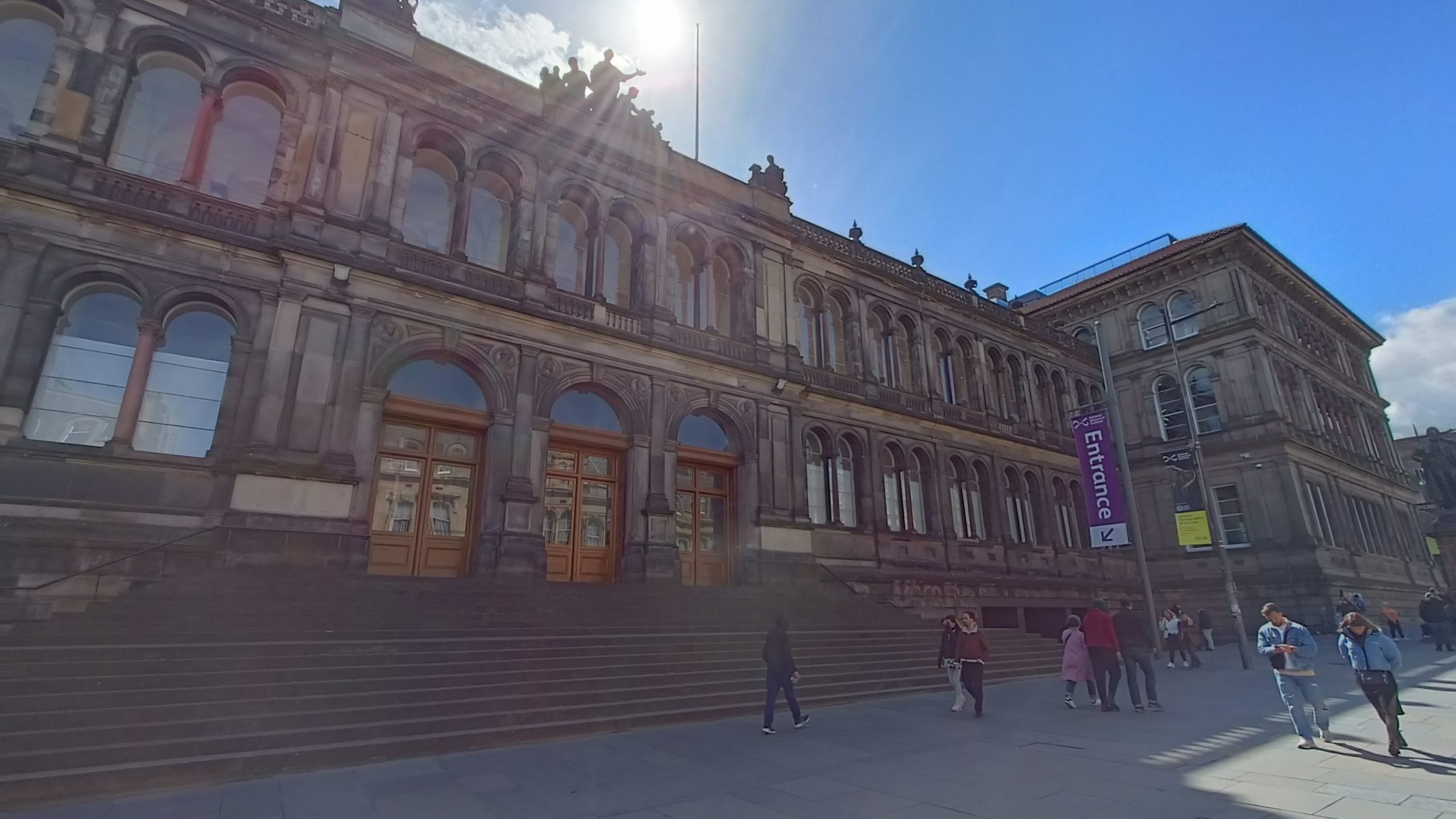 A historic stone building with arched windows and wide steps, where people gather under a bright blue sky—visit the National Museum of Scotland for an unforgettable experience.