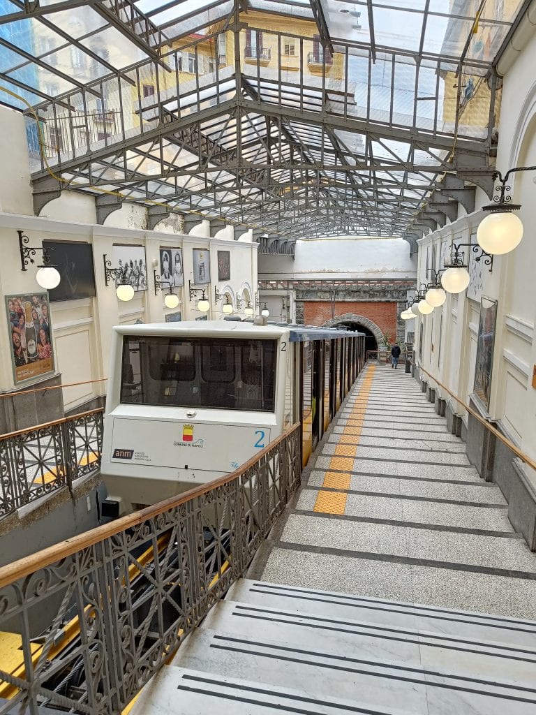 A funicular train at a covered station, with stairs and railings on both sides and bright hanging lights above—an iconic part of getting around Naples and experiencing unique Naples transportation.