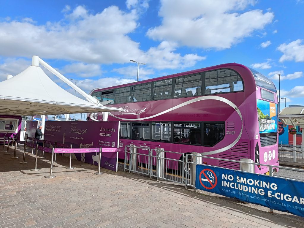 A purple double-decker bus at a station with a "No Smoking Including E-Cigarettes" sign in front highlights the clean and efficient transport in Glasgow, making getting around Glasgow easy and comfortable for everyone.