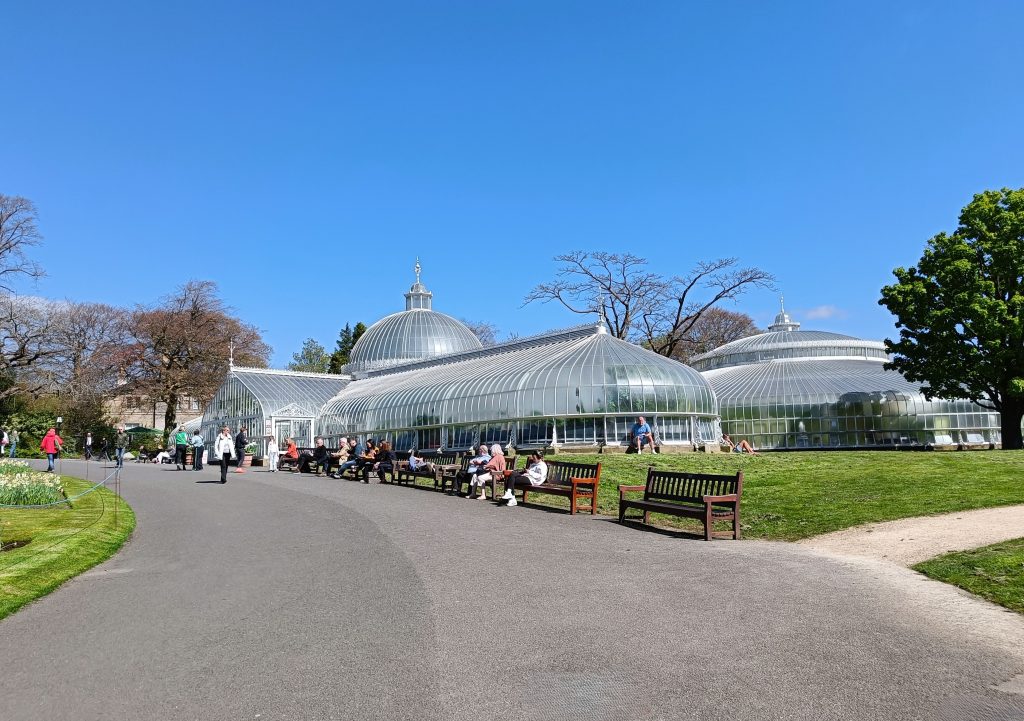 People sit on benches near a large glass greenhouse in a park under a clear blue sky—one of the 10x Best things to do in Glasgow for those seeking relaxation and natural beauty.