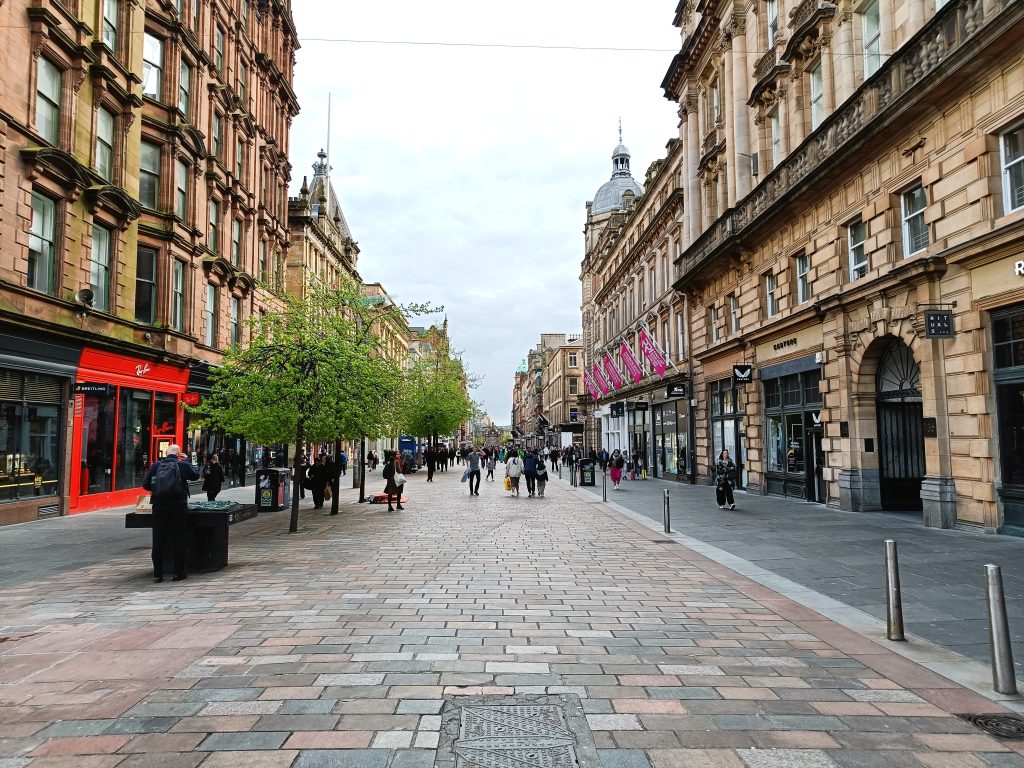 A pedestrian street lined with shops, trees, people walking, and historic buildings on both sides—truly one of the 10x Best things to do in Glasgow.