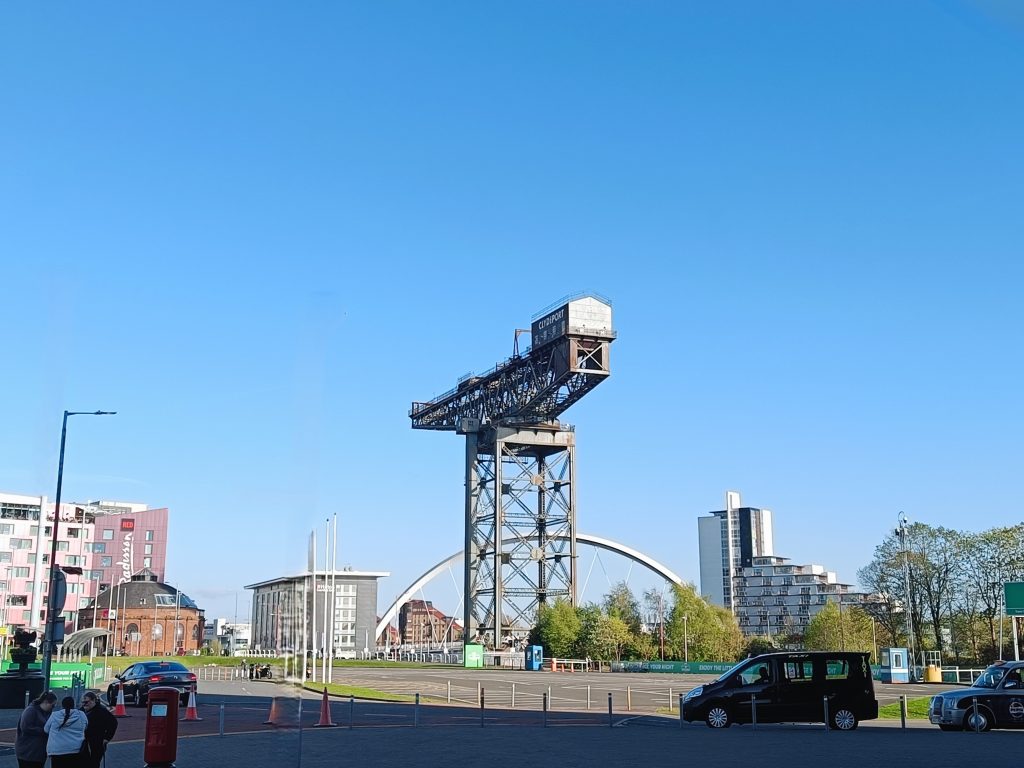 A large crane structure stands tall against a clear blue sky in an urban area with cars and people nearby—an iconic sight often featured on "10x Best things to do in Glasgow" lists.